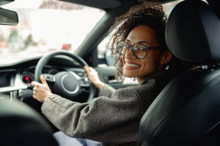 A woman smiling and wearing glasses, looking over her shoulder in the driver's seat of a car